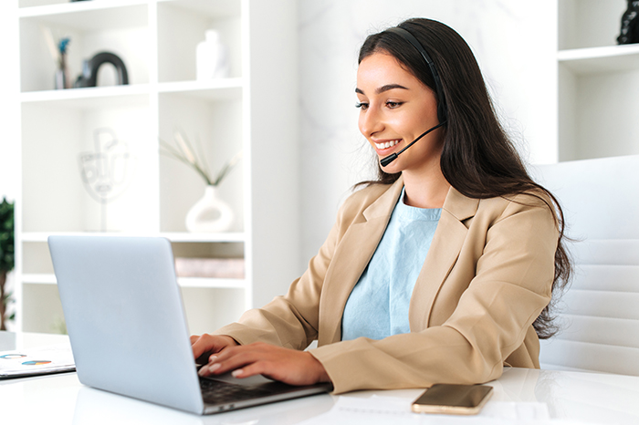 Friendly positive woman, call center operator with headset, working on support hotline in a modern office with a laptop, looking at the screen, smiling. Call center, customer support service